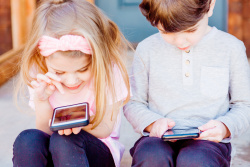 A young girl and boy sit side by side. Each of them has a smartphone in their hands. They are looking down at the phones.