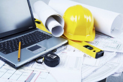 A laptop sits on top of plans with a hard hat and other gear